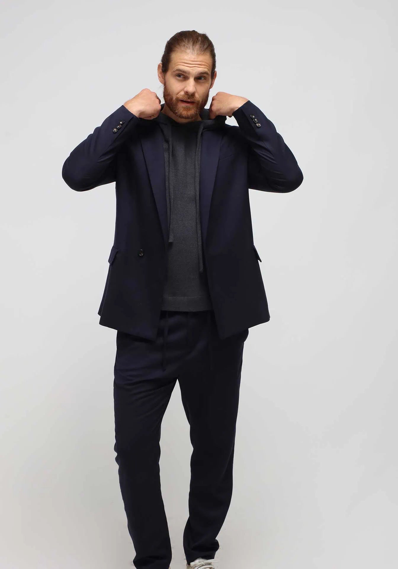A man wearing a blue cupro double-breasted single button jacket and comfortable traveller pants with a cashmere hoodie by Humans & Land on a white seamless background