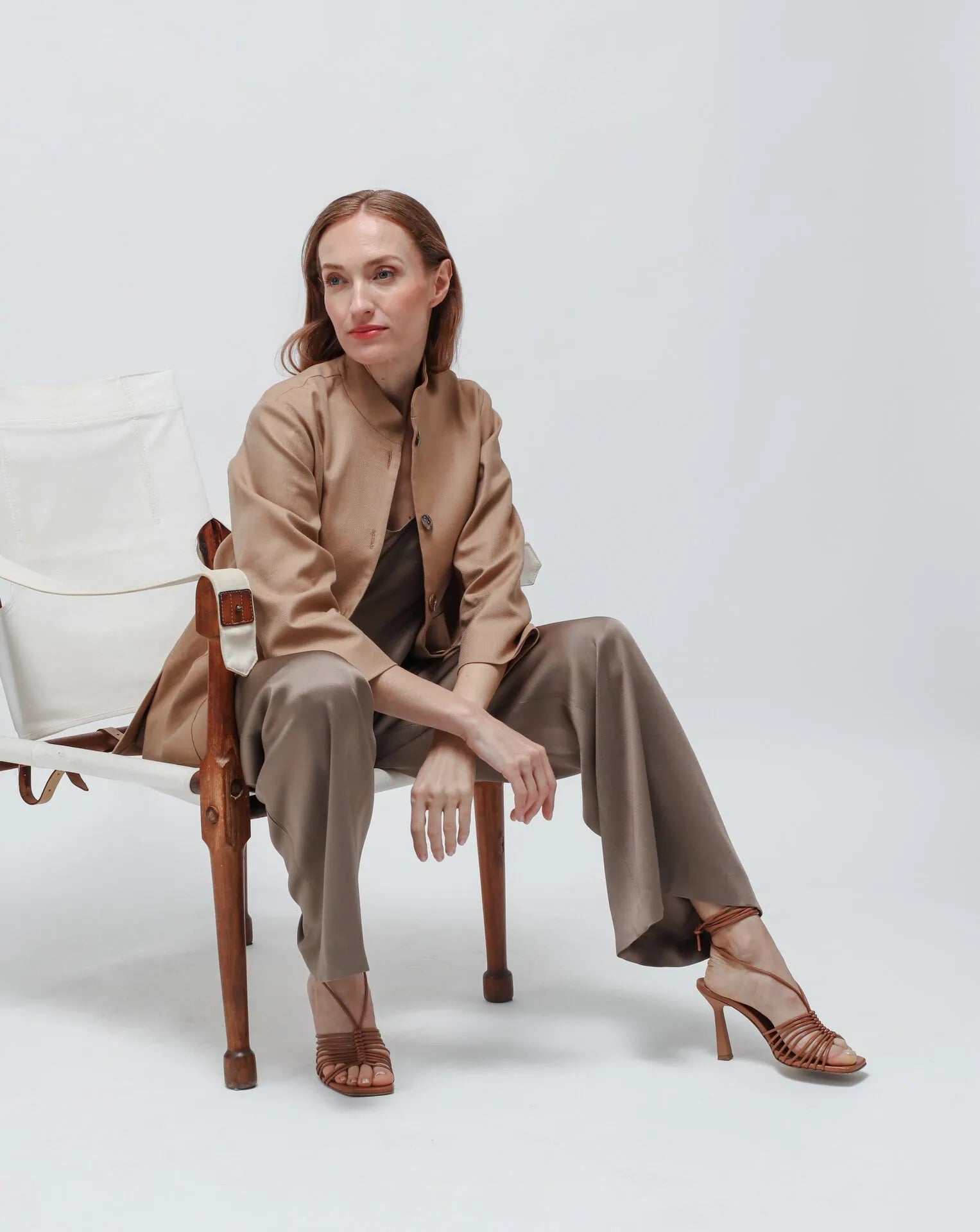 A woman wearing a Korean collar beige jacket with olive green satin pants sitting on a chair in a white seamless background.