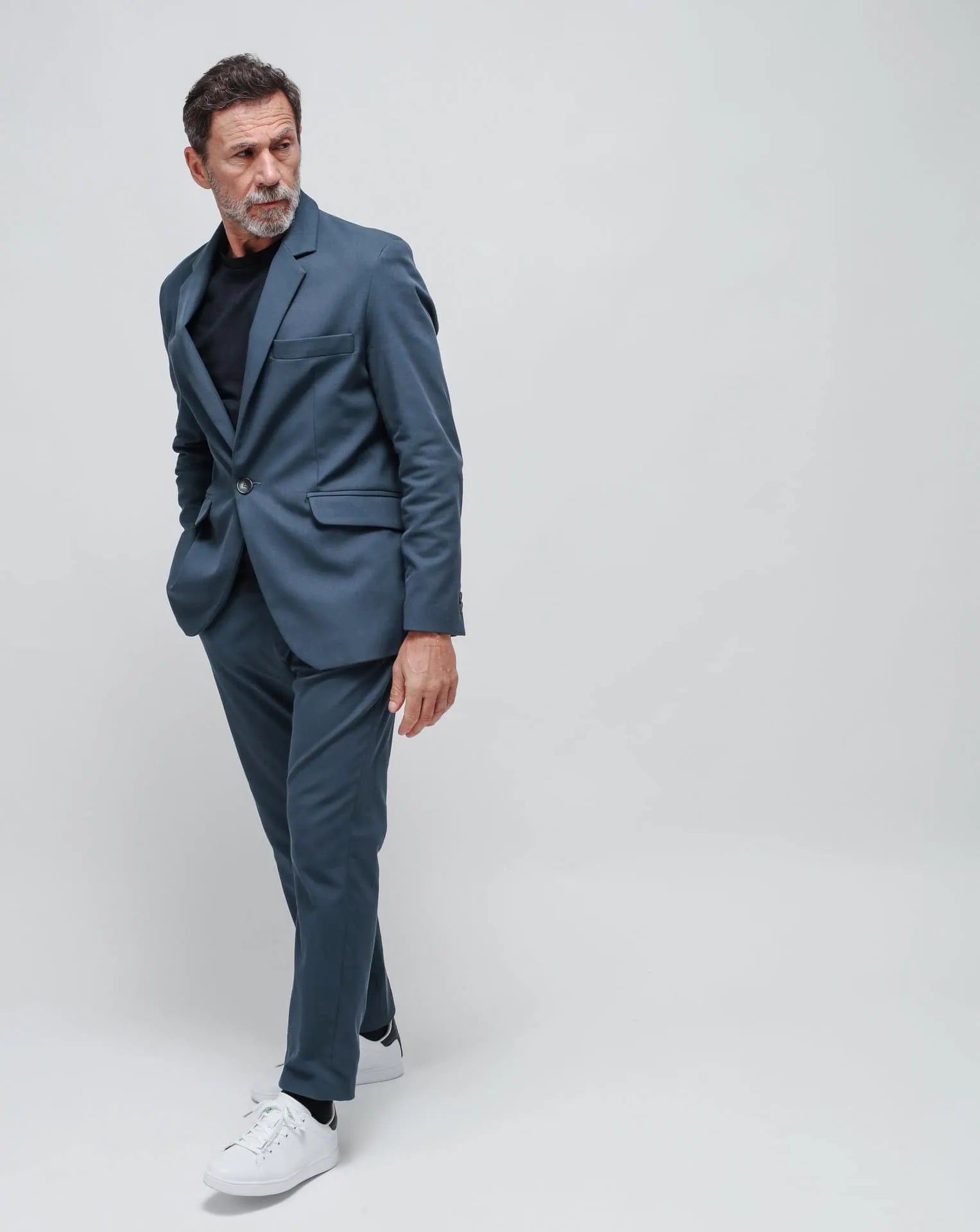 A man wearing a blue one-button summer jacket and a blue t-shirt, standing against a white seamless background.