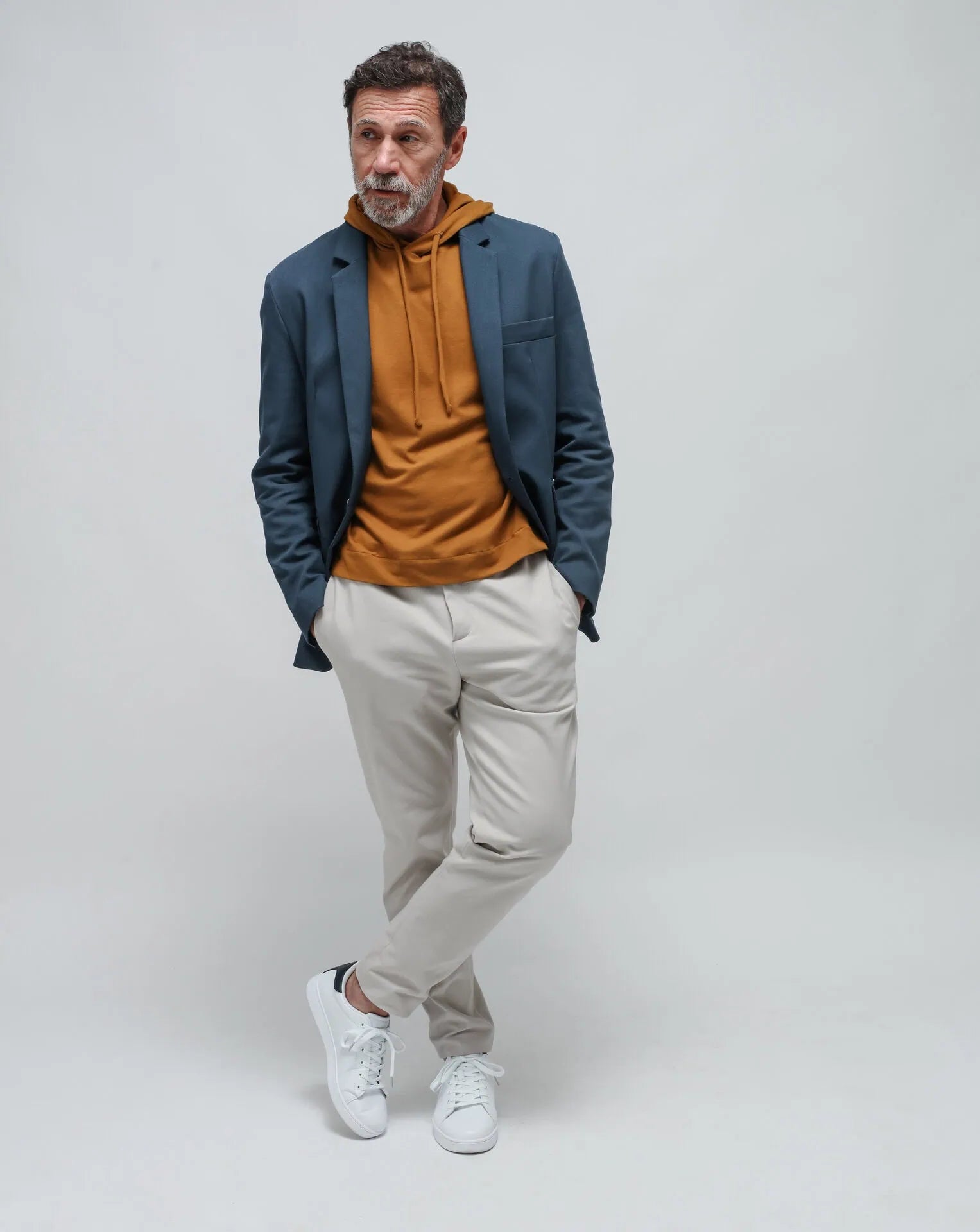 A man wearing a blue one-button summer jacket and a okra hoodie, standing against a white seamless background.