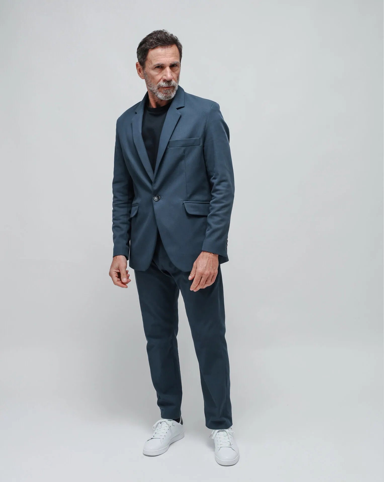 A man wearing a blue one-button summer jacket and a blue t-shirt, standing against a white seamless background.