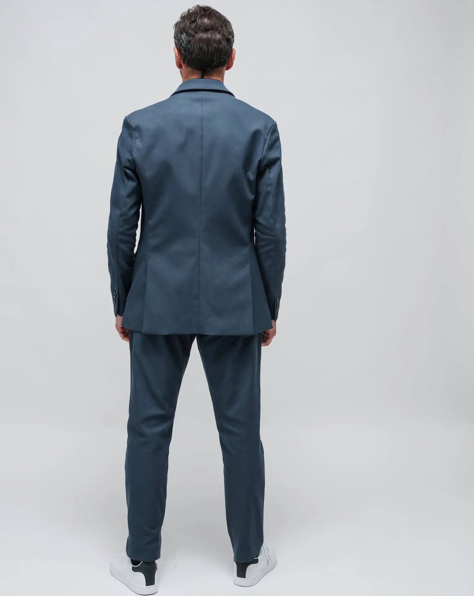 A man wearing a blue one-button summer jacket and a blue t-shirt, standing against a white seamless background.