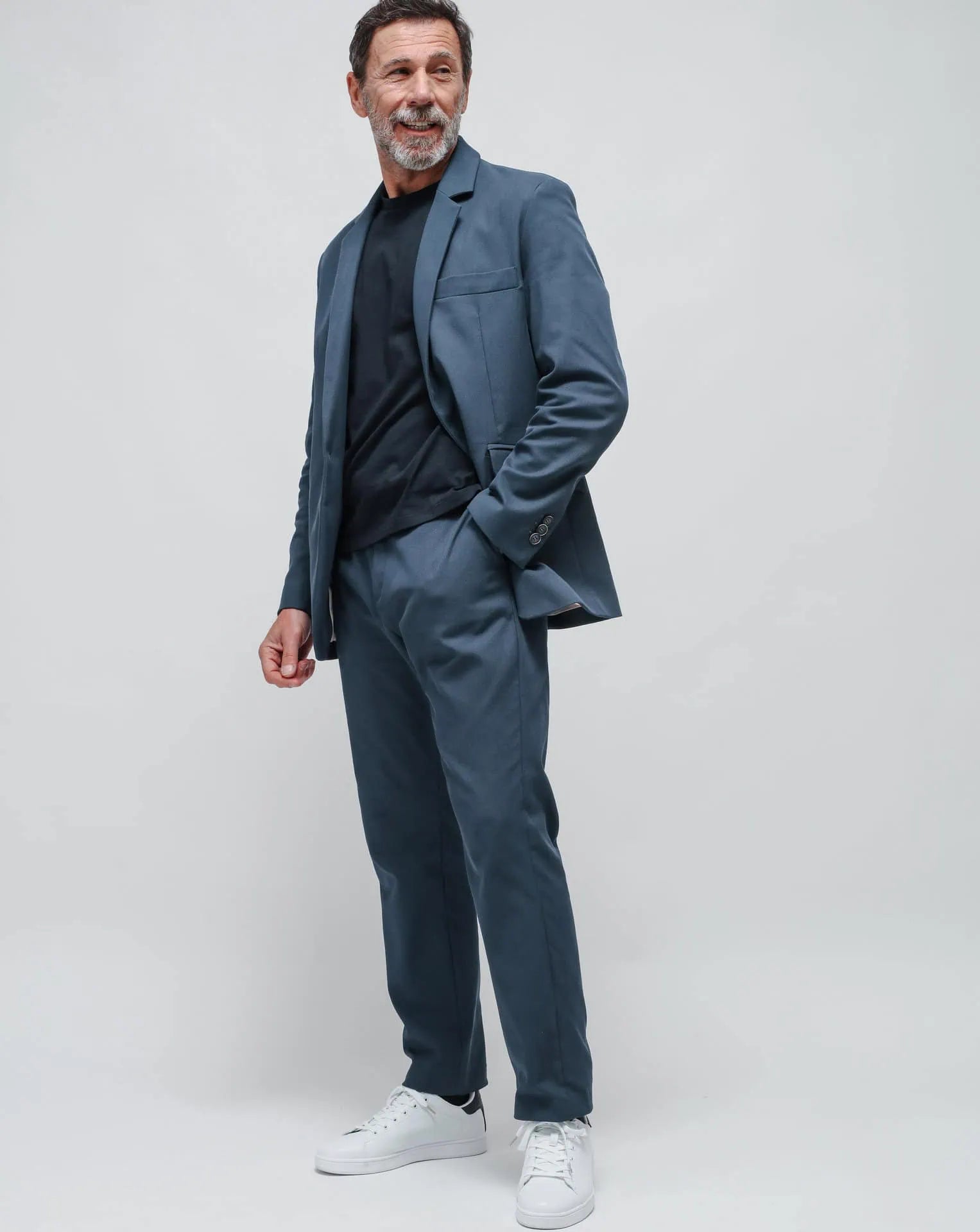 A man wearing a blue one-button summer jacket and a blue t-shirt, standing against a white seamless background.