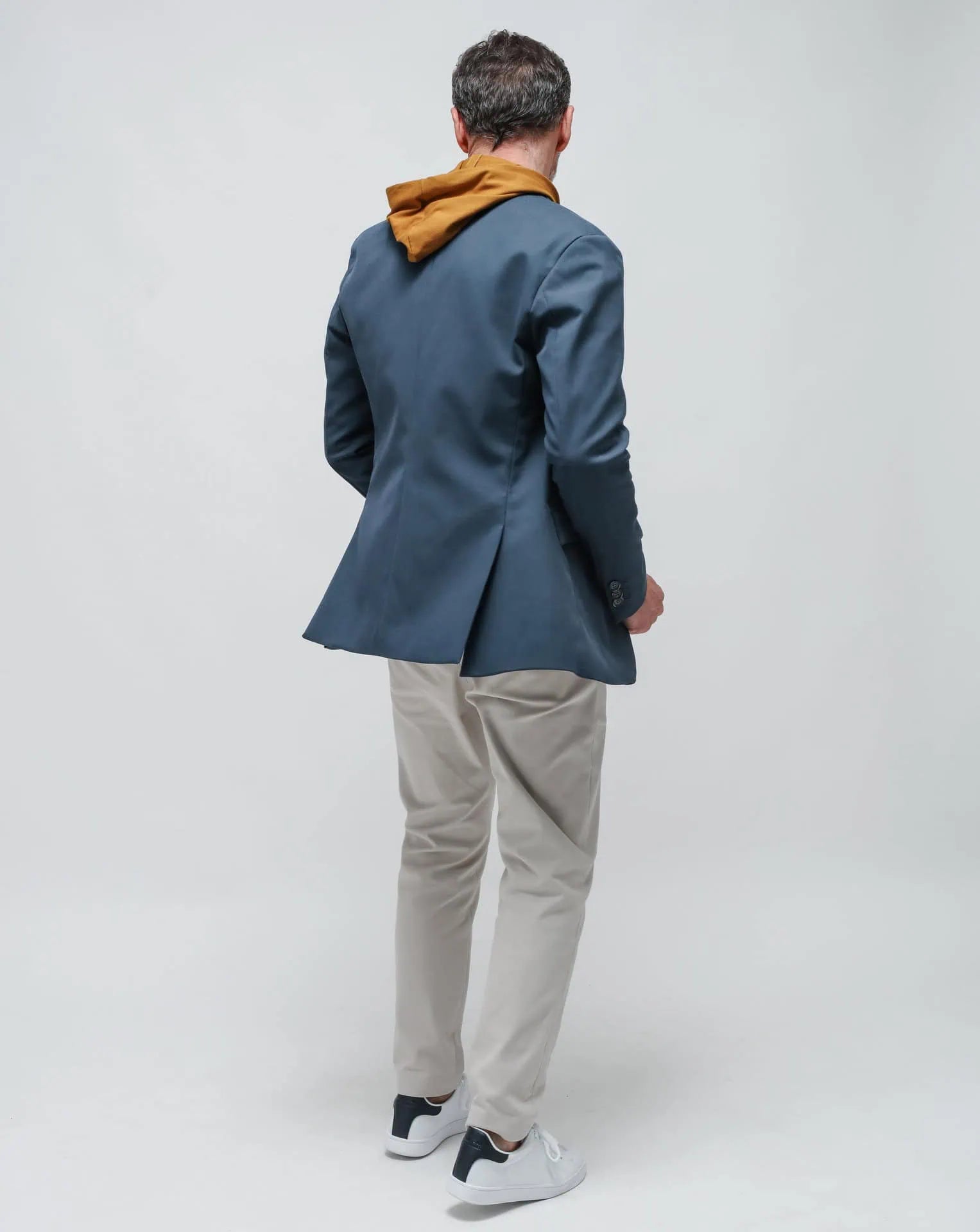 A man wearing a blue one-button summer jacket and a okra hoodie, standing against a white seamless background.