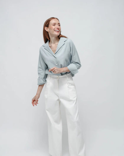 A women in white background wearing a light blue bowling shirt and white Punto Milano palazzo pants.