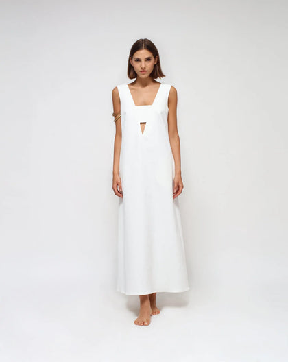 A woman standing in a studio setting, wearing a white, V-neck, ankle-length dress with a side zipper and side pockets.
