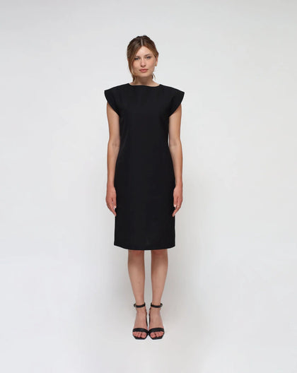 A woman standing against a plain background, wearing a midi black sheath dress with short sleeves and a round neckline.
