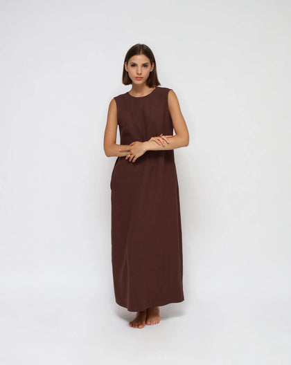 A woman standing, wearing a long, brown, linen, sleeveless, crew neck women's dress with open V-back and side pockets.