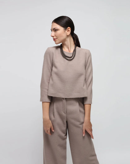 A female model wearing a beige wool flared top with crewneck and buttons down the back, paired with matching pants.