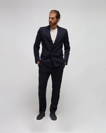 A man standing with hands in pockets, wearing a blue double-breasted suit with a beige t-shirt underneath.