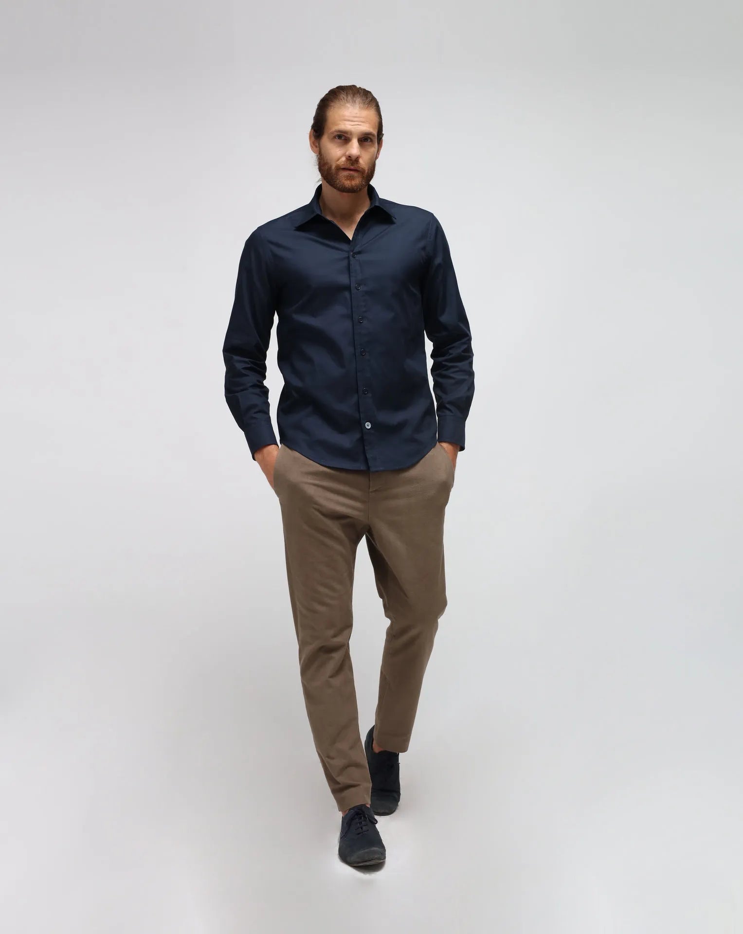 A man wearing a cotton blue shirt with brown pants standing in white seamless background.