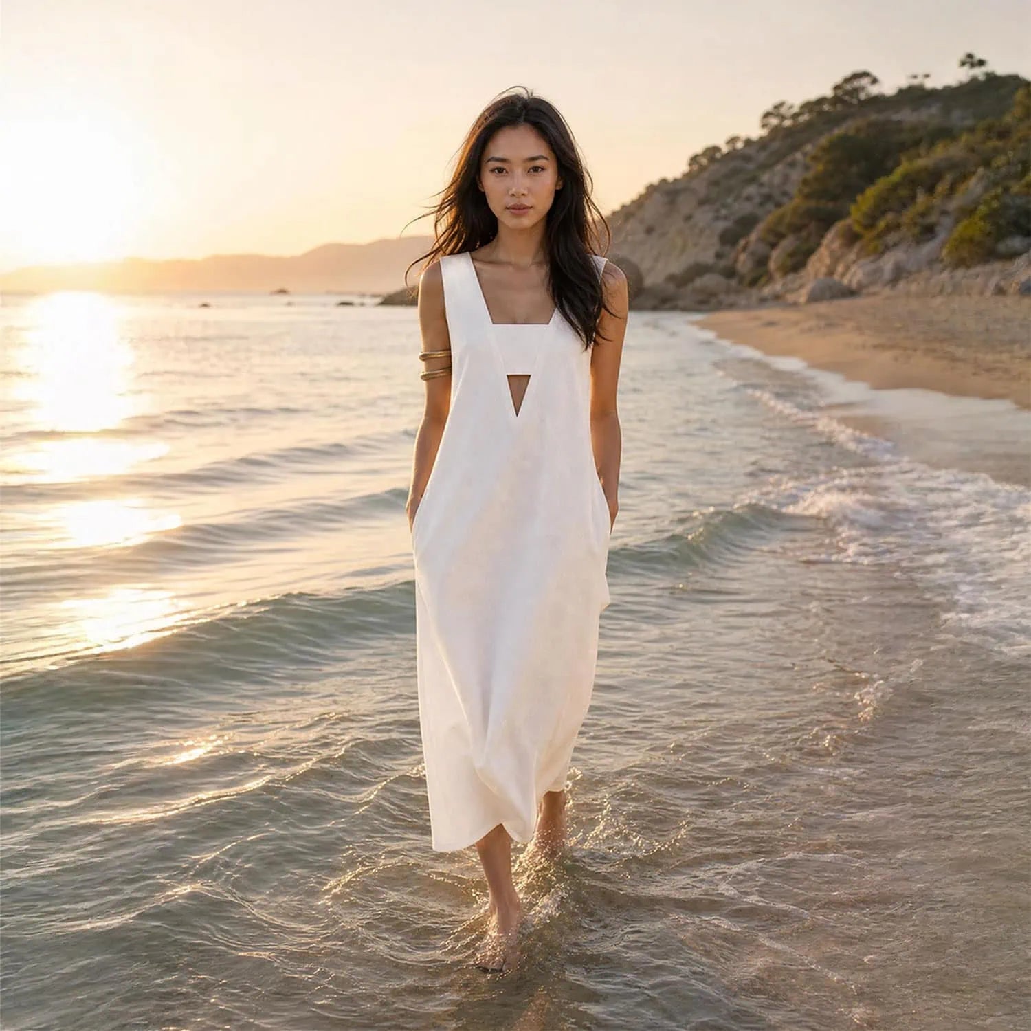 An Asian woman wearing a white long maxi dress is walking on a beach with her feet in the water looking into camera