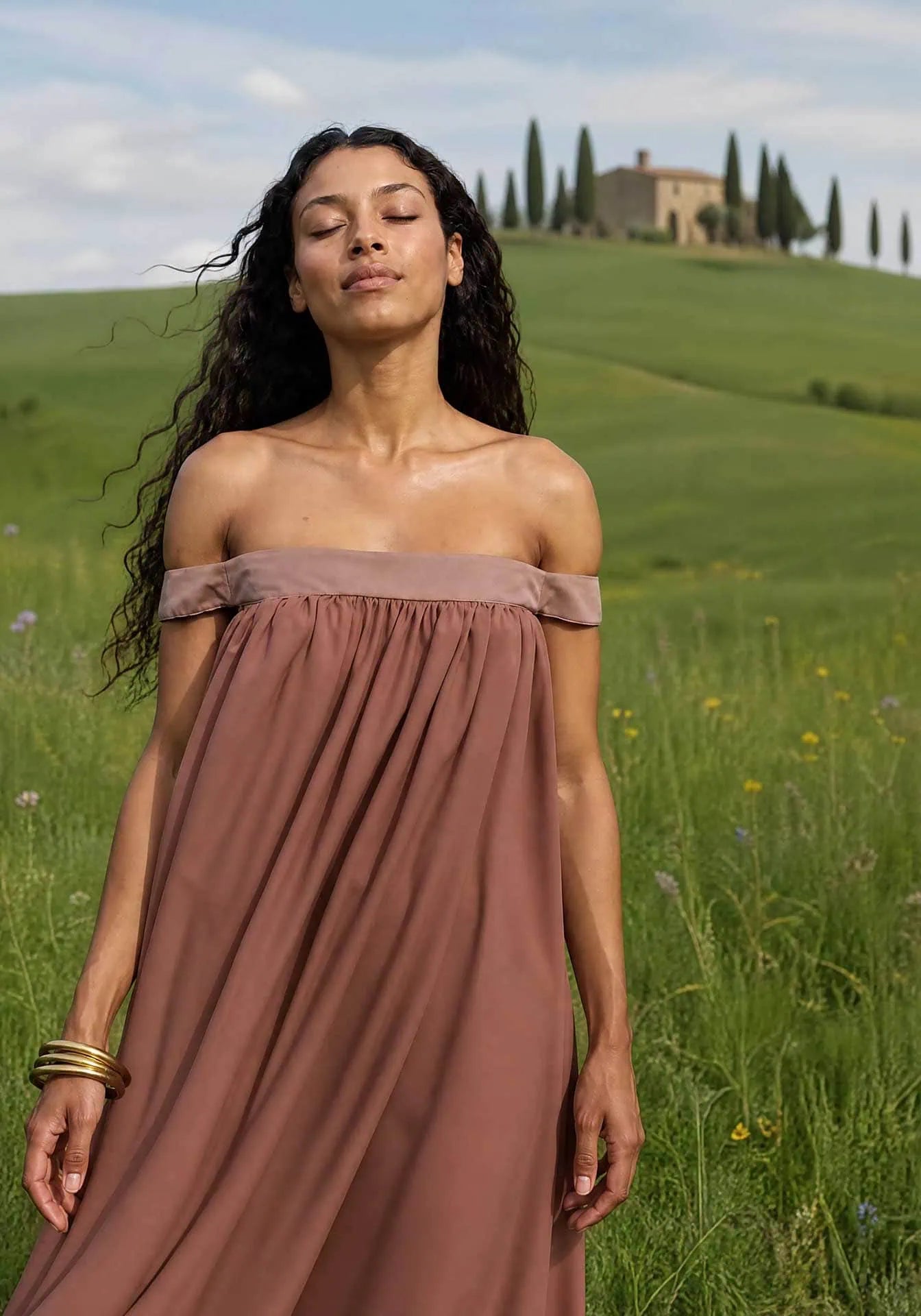 A woman wearing a brown silk strapless dress is standing on a green field with an Italian country house far in the background