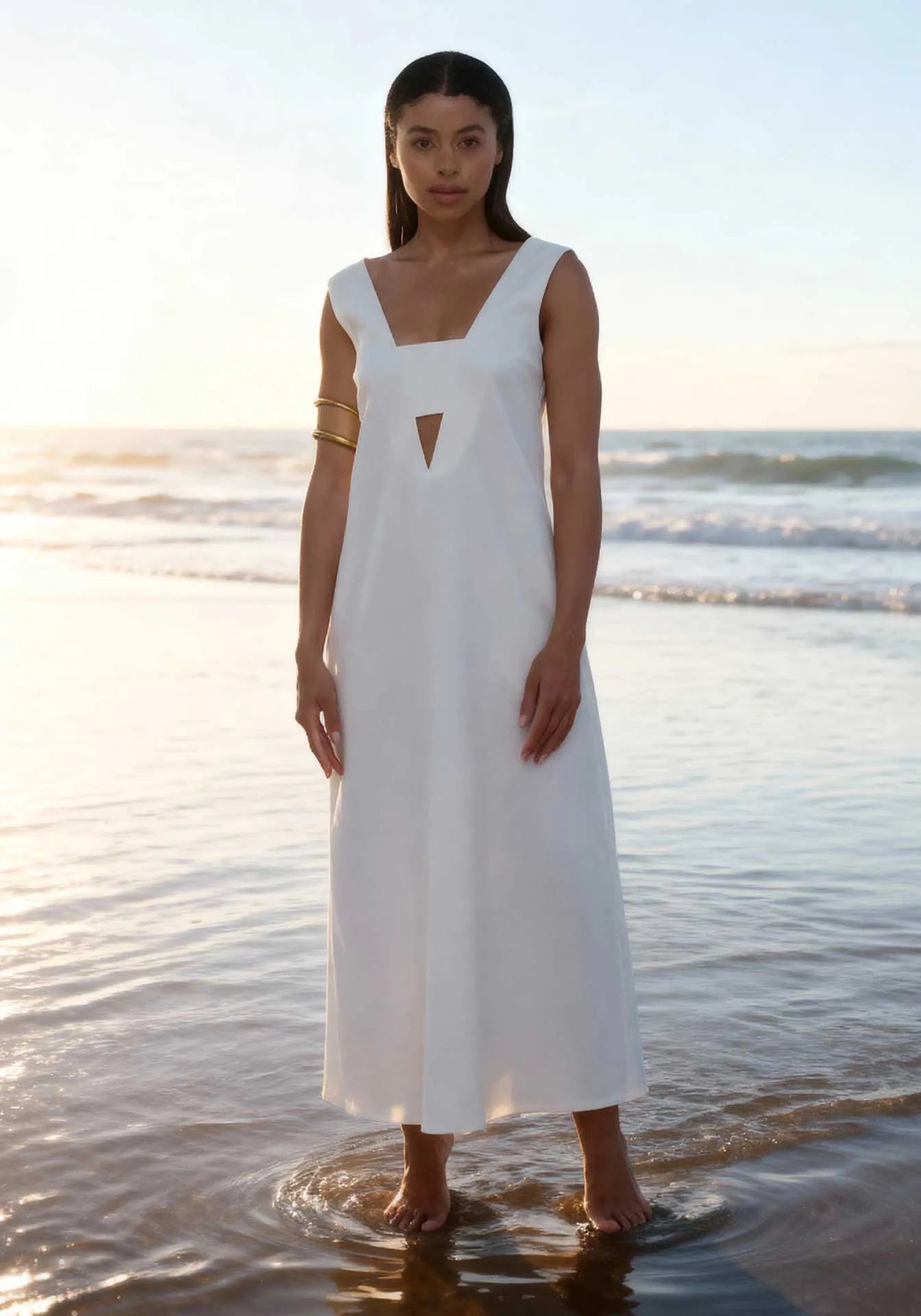A woman with her feet in the sand and ocean wearing a white long summer midi dress by Humans & Land