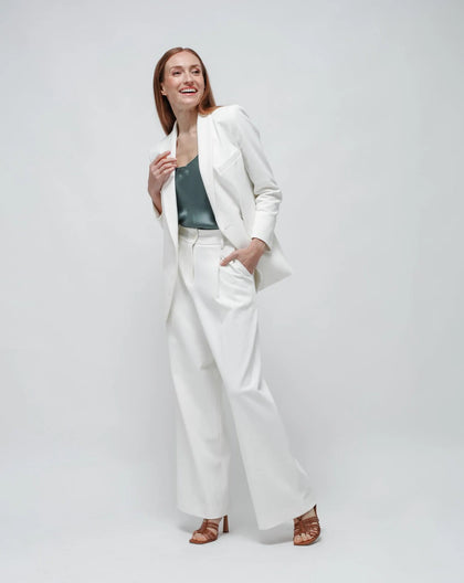 A model wearing a white, double-breasted, oversized jacket with a shawl lapel and flap pockets, paired with white trousers.