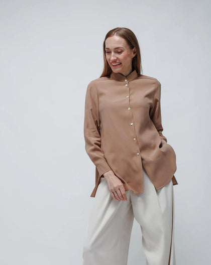 A woman standing, wearing a beige long Korean cupro shirt with a flared design and a necktie, paired with white trousers.