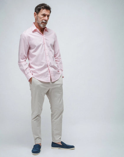 A man wearing a pink, long-sleeved,  shirt with simple lines, paired with beige pants and blue shoes, standing against a white seamless background.