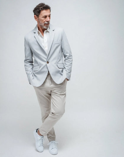 A man wearing a sky blue one-button summer jacket and beige trousers, standing with one hand in a pocket, against a white background.