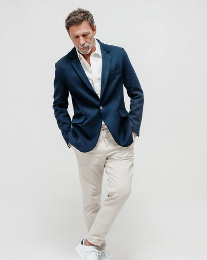 A man wearing a navy blue one-button jacket in Punto Milano (Milano Stitch) and light beige trousers, standing  a white surface.