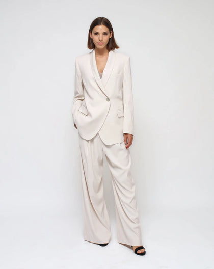 A female model wearing a white tailleur, consisting of a white-button jacket and palazzo pants, standing against a plain background.