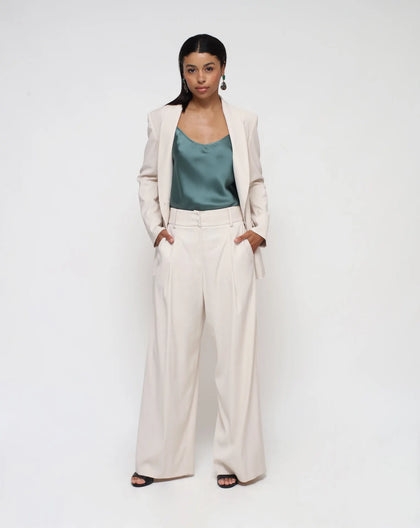 A model wearing a white double-breasted jacket with a shawl lapel and a single button. The jacket has pockets with flaps and is paired with a green top and beige trousers.