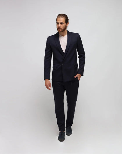 A man wearing a blue double-breasted one button jacket and blue trousers standing against a white background.