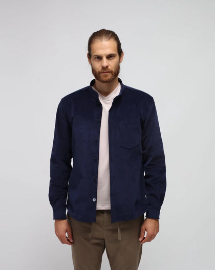 A man wearing a navy blue Korean collar shirt-jacket with a classic cuff and applied pocket, paired with a beige colored t-shirt and beige trousers.
