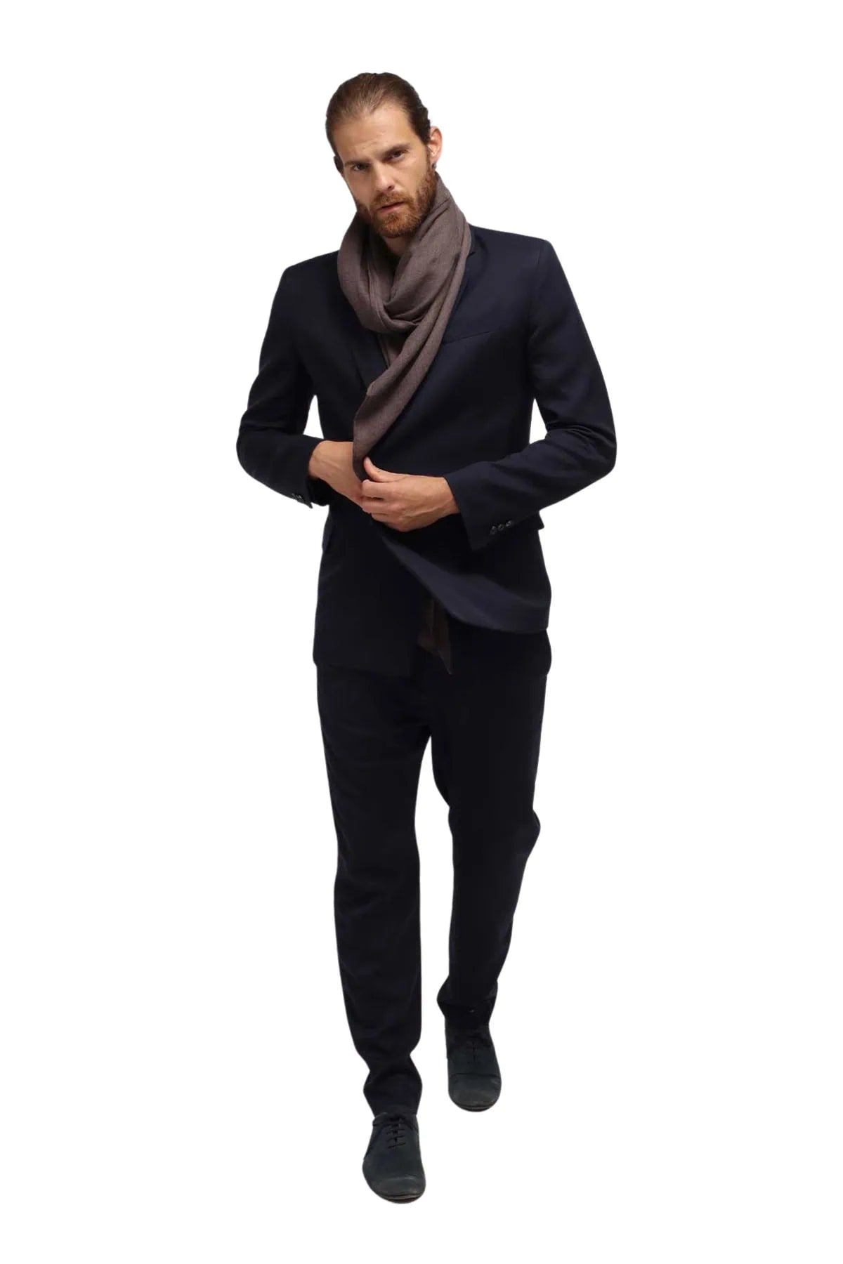 A man wearing a dark navy suit, a brown scarf, and black shoes walking against a solid white background.