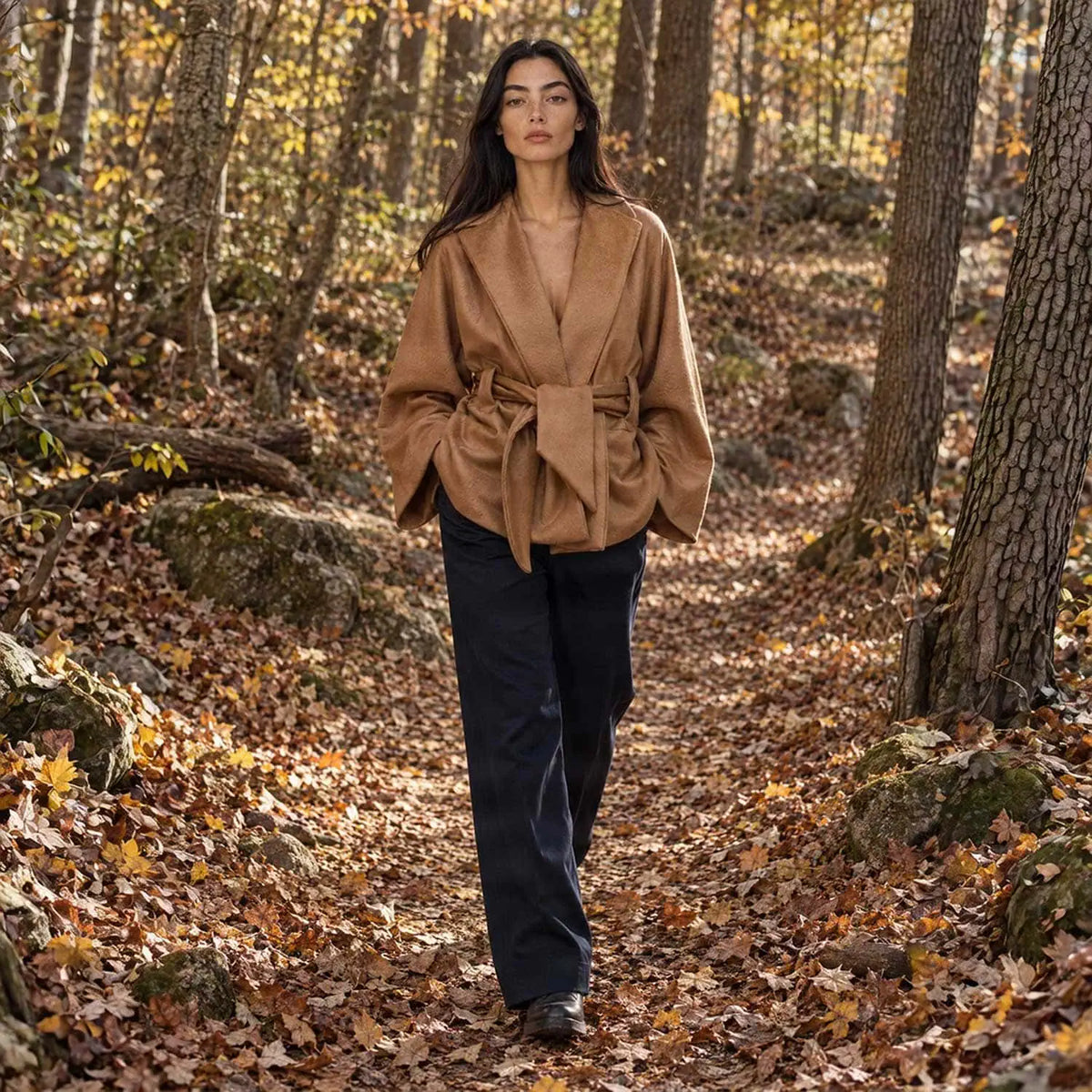A woman walks on a forest path covered in fallen autumn leaves, wearing a camel-brown belted wrap kimono jacket and dark blue wide-leg trousers.