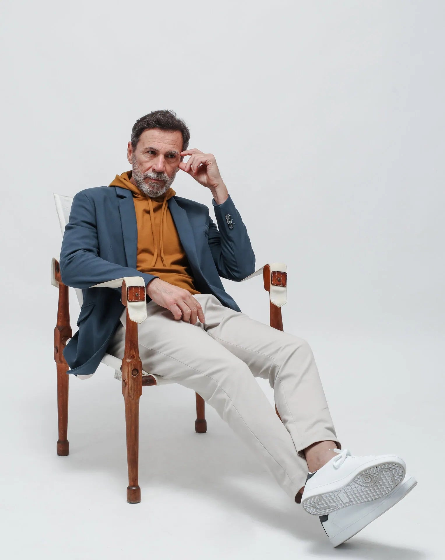 Man sitting on a chair wearing a blue blazer, mustard sweater, beige pants, and white sneakers.